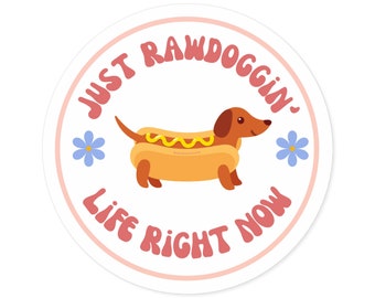 May include: A white circle sticker with a pink border. The sticker features a cartoon dachshund dressed as a hot dog. The text "Just Rawdoggin' Life Right Now" is written in a pink, retro font around the dachshund.