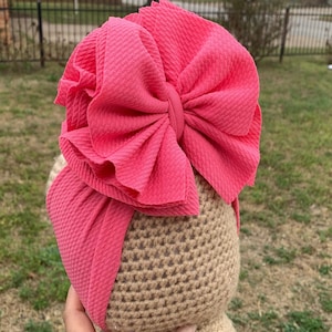 May include: A large, bright pink bow made of fabric with a textured surface. The bow is tied around a brown crocheted headband.