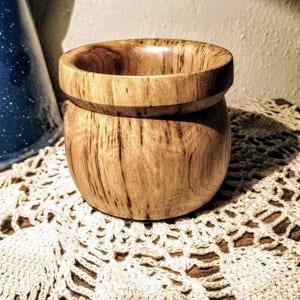 Puede incluir: Un pequeño cuenco de madera redondo con fondo redondeado y borde ligeramente acampanado. El cuenco está hecho de madera marrón claro con vetas marrones más oscuras.