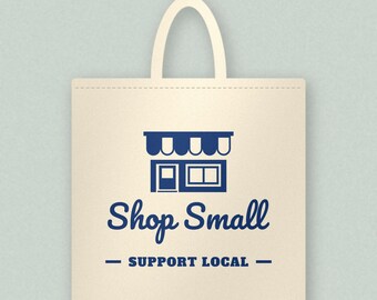 May include: A beige canvas tote bag with two handles featuring a dark blue graphic of a storefront with an awning. Below the graphic, the text reads Shop Small in a script font and SUPPORT LOCAL in a bold, sans-serif font.
