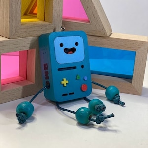 Puede incluir: Un robot de juguete de madera azul verdoso con una cara sonriente, un diseño de mando de juego y el texto "BMO" en el lateral. El robot tiene cuatro brazos de cuentas de madera azul verdoso.