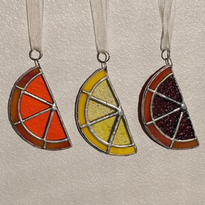 Op de afbeelding: Drie glas-in-lood ornamenten in de vorm van sinaasappelschijfjes. De ornamenten zijn rood, geel en paars. Elk ornament heeft een wit lint aan de bovenkant.