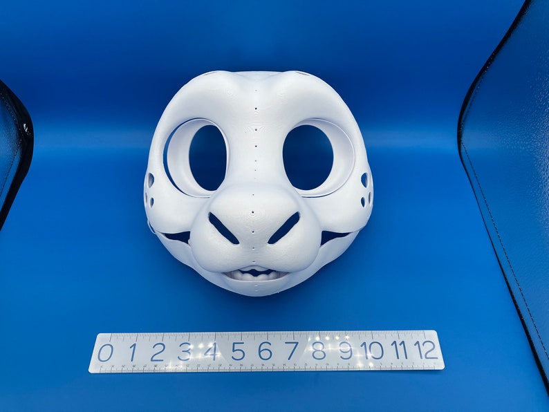 K&ouml;nnte beinhalten: Wei&szlig;e, unfertige Felltiermaske mit gro&szlig;en Augenl&ouml;chern und einer kleinen Nase. Die Maske liegt auf einer blauen Oberfl&auml;che mit einem Lineal, das von 0 bis 12 markiert ist.
