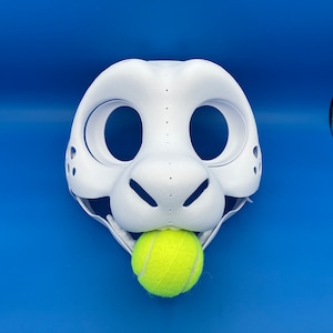 K&ouml;nnte beinhalten: Wei&szlig;e pelzige Tiermaske mit gro&szlig;en Augenl&ouml;chern und einer kleinen Nase. Die Maske ist offen und hat einen gelben Tennisball im Maul.