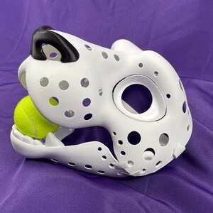 May include: White dalmatian dog mask with black nose and holes throughout. A yellow tennis ball is in the mouth of the mask.