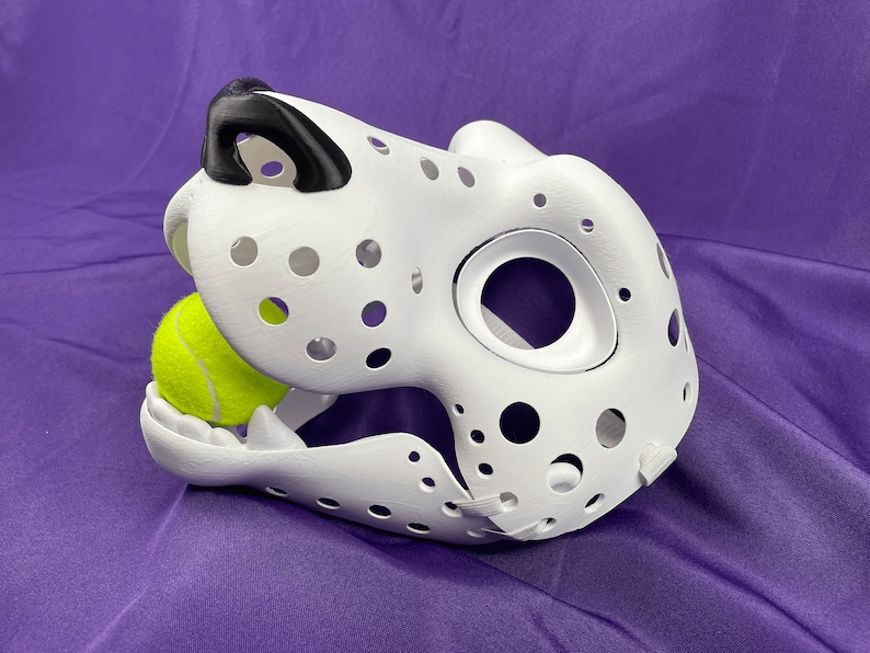 May include: White dalmatian dog mask with black nose and holes throughout. A yellow tennis ball is in the mouth of the mask.