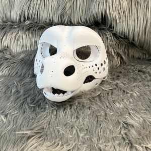 May include: White unpainted furry animal head mask with holes for eyes and nose. The mask is on a gray fluffy surface.