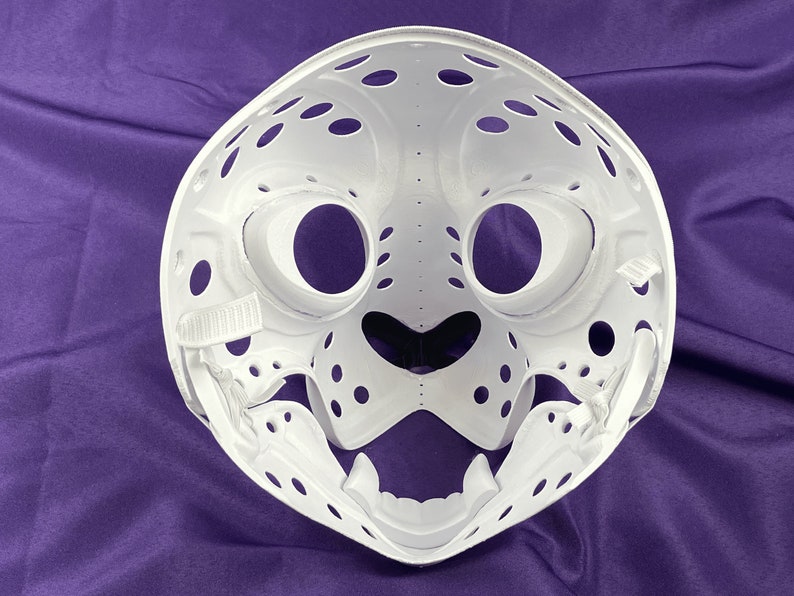 May include: White plastic mask with a detailed design resembling a cat's face. The mask has large eye holes, a small nose, and a wide mouth with teeth.