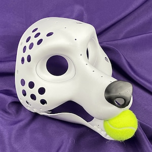 Op de afbeelding: Witte, onafgewerkte hondenkop voor een fursuit met een zwarte neus en een gele tennisbal in de mond. De kop heeft veel kleine gaten voor ventilatie.