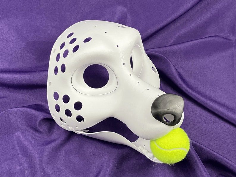 Op de afbeelding: Witte, onafgewerkte hondenkop voor een fursuit met een zwarte neus en een gele tennisbal in de mond. De kop heeft veel kleine gaten voor ventilatie.