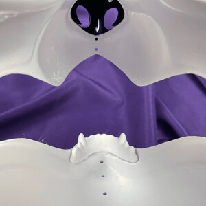 Op de afbeelding: Wit plastic masker met een grote open mond en een klein neusgat. Het masker ligt op een paarse stoffen achtergrond.
