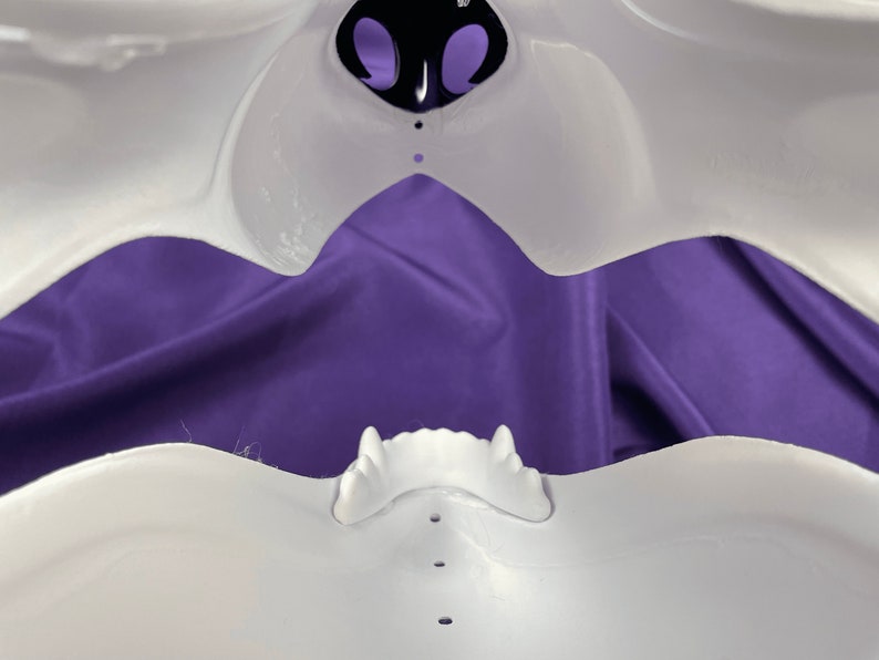 Op de afbeelding: Wit plastic masker met een grote open mond en een klein neusgat. Het masker ligt op een paarse stoffen achtergrond.