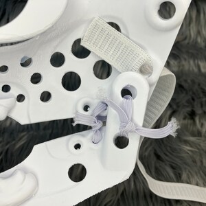 May include: White plastic helmet with a white elastic strap and a white velcro strap. The helmet has a series of holes in the plastic.