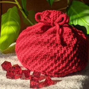 Puede incluir: Bolsa de dados de crochet roja con cierre de cordón. La bolsa está llena de dados rojos.