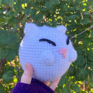 May include: A white crocheted plush toy shaped like a cat with black eyes and a pink nose. The toy is being held in a hand wearing a purple sweater.