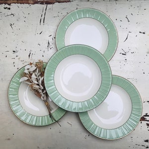 Op de afbeelding: Vier witte keramische borden met een mintgroen en goudkleurig randontwerp. De borden zijn gerangschikt op een verweerd wit oppervlak, met een kleine bundel gedroogde bloemen. De borden hebben een geribbeld patroon op de rand.