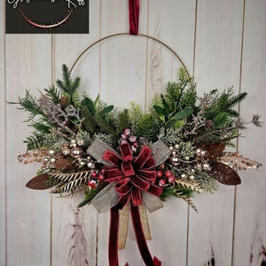 May include: A gold metal wreath with a burgundy velvet bow and gold ribbon. The wreath is decorated with greenery, berries, and gold accents. The wreath is hanging on a white wooden wall.