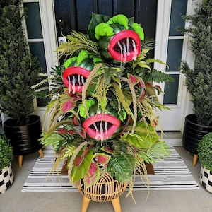 May include: A whimsical Halloween decoration featuring a woven basket planter with a plant arrangement. The arrangement includes three large, green, open-mouthed figures with red lips and white drool, surrounded by various green and red foliage. The text "Glitter in the Ruff" is at the bottom.