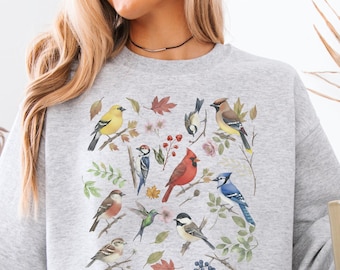May include: A light gray sweatshirt featuring a colorful illustration of various birds, leaves, and flowers. The design includes a red cardinal, blue jay, and other birds. The sweatshirt is made of a soft material and is suitable for casual wear.