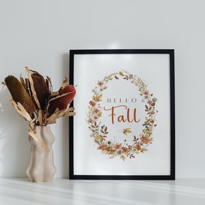May include: A black framed print with a watercolor wreath of fall leaves, flowers, and mushrooms. The text "HELLO FALL" is in the center of the wreath. The print is sitting on a white surface next to a vase with dried brown and red flowers.