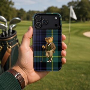 Könnte beinhalten: Smartphone-Hülle mit blauem, grünem und gelbem Tartanmuster. Sie zeigt einen Cartoon-Bären in Golfkleidung, der einen Golfschläger hält. Im Hintergrund ein Golfplatz mit Fahne und Golfschlägern.