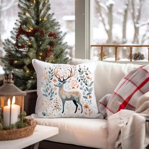 May include: A white throw pillow with a floral pattern featuring a deer with antlers. The deer is surrounded by leaves, flowers, and berries in shades of blue, green, orange, and pink.