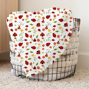 May include: A white blanket with a repeating pattern of red ladybugs, yellow flowers, and green leaves. The blanket is folded over a wire basket filled with white towels. The ladybugs have black spots and are set against a white background.