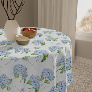 May include: A round white tablecloth with a watercolour pattern of blue hydrangea flowers and green leaves. The tablecloth is draped over a round table. A white vase, a small basket of apples, and two bowls are on the table.
