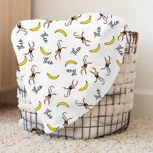 May include: White baby blanket with a repeating pattern of monkeys and bananas. The name "Theo" is written in black cursive throughout the blanket.