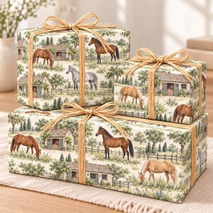 May include: Three rectangular gift boxes wrapped in cream-colored paper featuring a repeating pattern of horses in a pasture with stables, trees, and fences. Each box is tied with a natural tan raffia ribbon tied in a bow on the top center.
