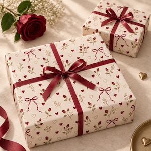 May include: Two square gift boxes wrapped in cream-colored paper featuring a repeating pattern of small red roses, green sprigs, red hearts, and red ribbon bows. Each box is tied with a smooth, dark red satin ribbon centered in a bow.