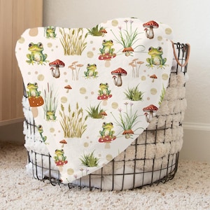 May include: A white blanket with a whimsical pattern of green frogs, red-capped mushrooms, and tall grass. The blanket is folded over a black wire basket with a fluffy, cream-colored lining. The design includes tan polka dots.