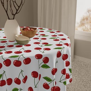 May include: A round tablecloth featuring a repeating pattern of red cherries with green leaves on a white background. The fabric drapes over a circular table, which holds a white vase with dark branches, a small woven basket, and a bowl.