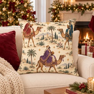 May include: A square throw pillow featuring a cream-colored fabric printed with a scene of three figures riding camels across a desert landscape. The figures wear robes in red, blue, and purple, and the design includes palm trees and stars.