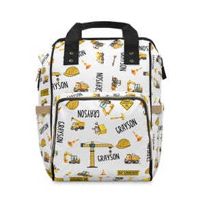 May include: White backpack with a construction theme. The bag features a black handle and straps, with a pattern of yellow construction vehicles, tools, and the name "Grayson". Includes side pockets.