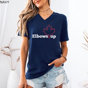 May include: A navy blue v-neck t-shirt with a red outline of a maple leaf and the text "Elbows Up" in white.