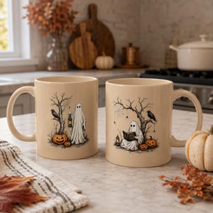 May include: Two beige ceramic mugs featuring black ink illustrations. The left mug shows a ghost holding a lantern near a tree, a crow, and pumpkins. The right mug depicts a ghost sitting under a tree reading a book, with a crow, spiders, and pumpkins.