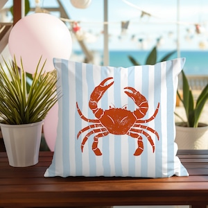 May include: A white throw pillow with blue stripes features a red crab illustration. The pillow is on a wooden surface.