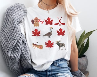May include: White t-shirt featuring a collection of Canadian-themed illustrations. The design includes red maple leaves, a beaver, a goose, hockey sticks, a canoe, and a moose. The shirt is made of a soft material.