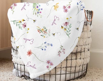 May include: White baby blanket with a floral pattern featuring various flowers, bees, and the name "Emilia" repeated four times.