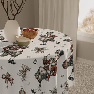 May include: A round tablecloth with a white background featuring a repeating pattern of toy drummers in red uniforms, holly sprigs, red bows, and musical notes. The fabric is draped over a circular table, with a white vase and a small woven bowl.