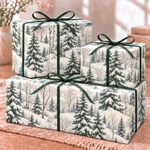 May include: Three rectangular gift boxes wrapped in white paper featuring a winter forest scene with snow-covered pine trees and birch trees. Each box is tied with a dark green satin ribbon tied in a bow on the top center.