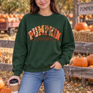 Peut inclure: Un sweat-shirt à manches longues vert foncé avec le texte PUMPKIN season écrit dans une police à blocs multicolores et à motifs. Le mot PUMPKIN est affiché en grandes lettres décoratives avec divers imprimés automnaux, et season en cursif.