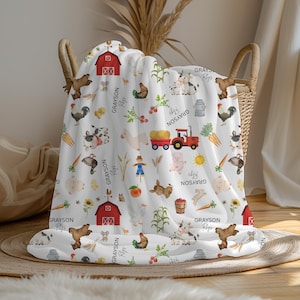 May include: White blanket with a farm animal pattern, including cows, pigs, chickens, and a red tractor. The blanket features a red barn, carrots, and the text "Grayson". The blanket is folded in a wicker basket.