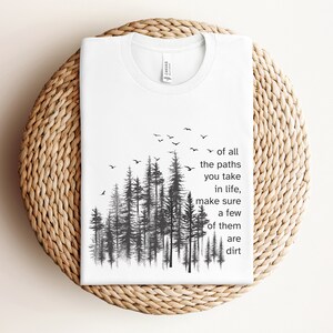 May include: White t-shirt with a black and white graphic of a forest with birds flying above. The text "of all the paths you take in life, make sure a few of them are dirt" is printed on the shirt.