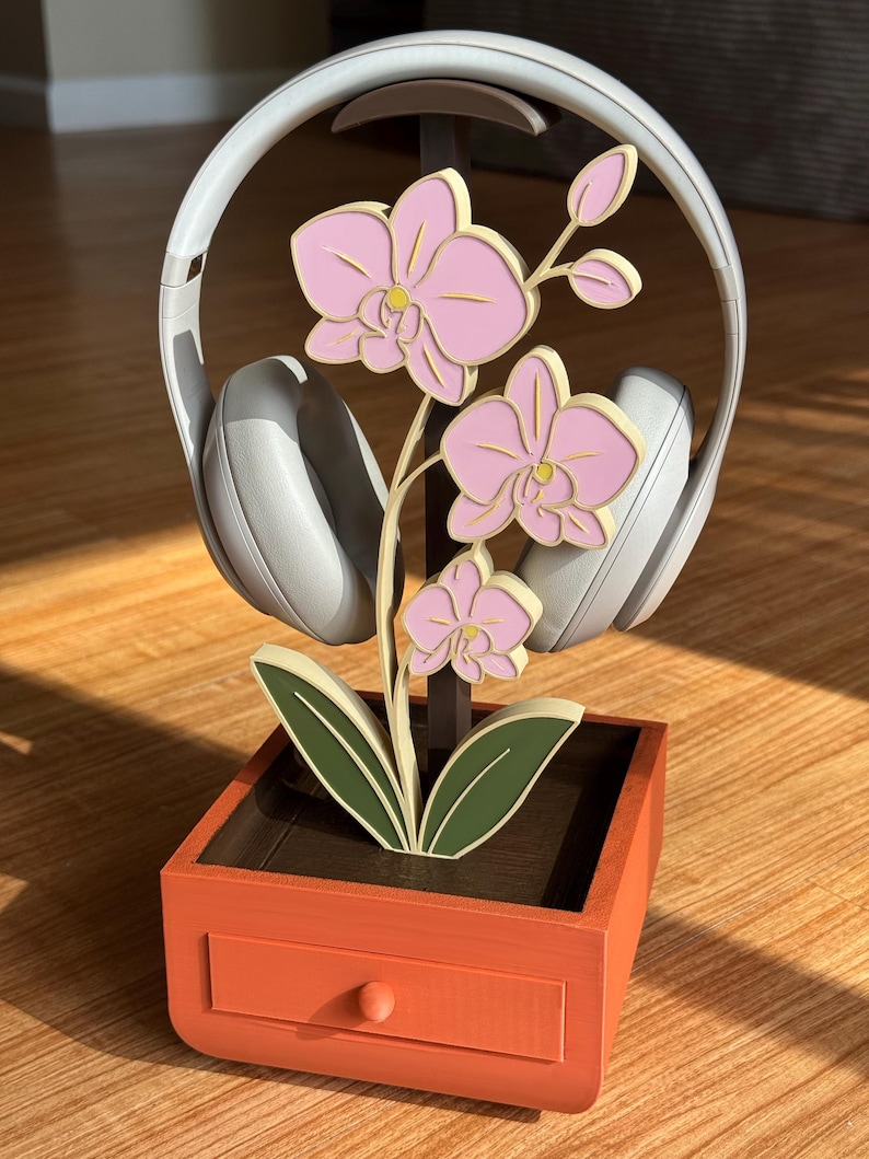 May include: A decorative headphone stand featuring a wooden orchid design with pink flowers and green leaves. The stand is orange with a small drawer and holds a pair of gray headphones. The stand is designed to hold headphones.