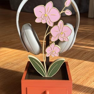 May include: A decorative headphone stand featuring a pink orchid design. The stand is made of wood and has a drawer. The headphones are white and rest on the stand. The stand is orange and brown.