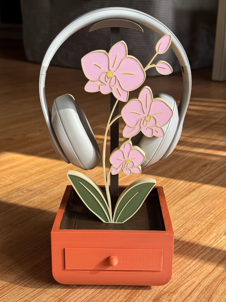 May include: A decorative headphone stand featuring a pink orchid design. The stand is made of wood and has a drawer. The headphones are white and rest on the stand. The stand is orange and brown.
