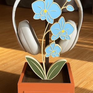 May include: A decorative headphone stand featuring a light blue orchid design. The stand has a brown stem and green leaves, set in an orange planter box with a small drawer. A pair of white headphones rests on the stand.