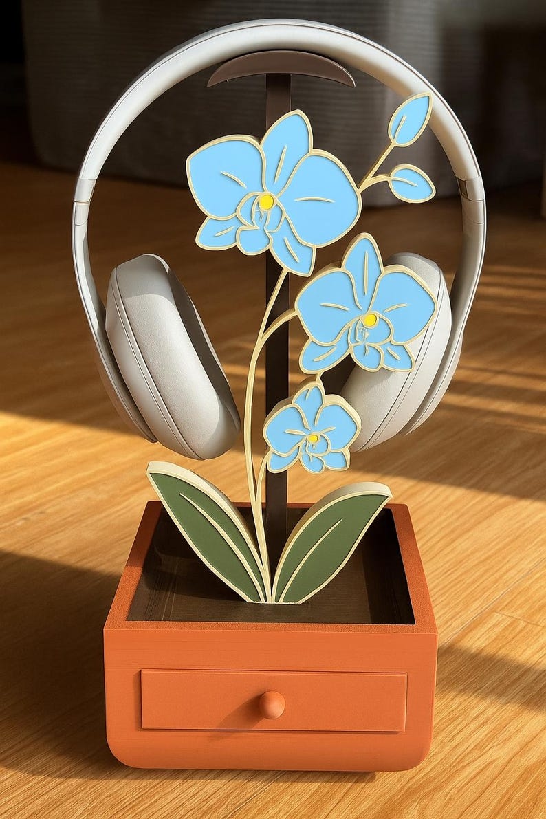 May include: A decorative headphone stand featuring a light blue orchid design. The stand has a brown stem and green leaves, set in an orange planter box with a small drawer. A pair of white headphones rests on the stand.